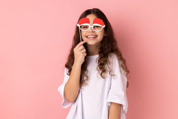 Portrait of little girl wearing white T-shirt holding funny paper glasses, having fun playing game, wearing masquerade accessory. Indoor studio shot isolated on pink background.