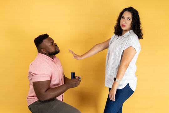 Portrait of young couple in casual clothing, man making a proposal to his girlfriend, woman refusing, showing stop palm gesture. Indoor studio shot isolated on yellow background.