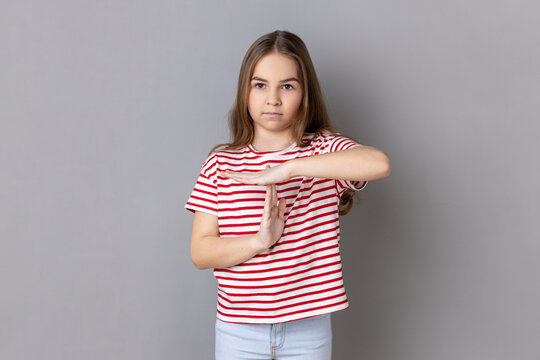 Portrait of frustrated little girl wearing striped T-shirt showing time out gesture, looking at camera, deadline with test in school. Indoor studio shot isolated on gray background.