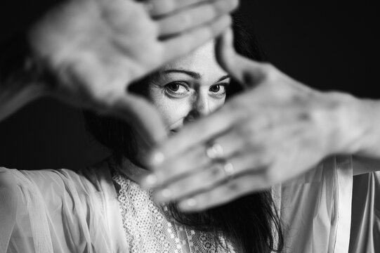 Anxiety, danger, protection - conceptual image of a woman covering her face with her hands, looking through them and peeking with one eye between her fingers on a dark studio background
