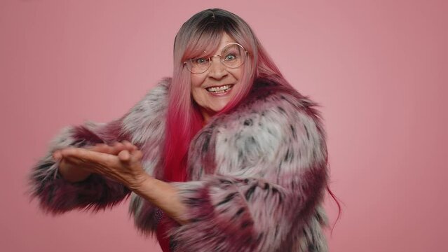 Happy rich stylish elderly woman showing wasting or throwing money around hand gesture, more tips dreaming about big profit body language. Senior grandmother posing isolated on pink studio background