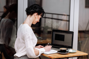 Young businesswoman sitting at table in cafe and writing in notebook. On table is laptop, cup of coffee and some sweet. Freelancer working in coffee shop. Student learning online. Business concept