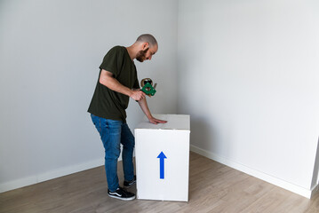 Un hombre joven usando una herramienta para precintar una caja de cartón blanca con cinta adhesiva...