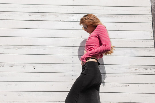 Beautiful Sexy Fashion Slender Girl With Trendy Sunglasses In A Fashionable Pink Knit Top With Long Sleeves And A Black Skirt Against A White Vintage Wooden Wall In Sunlight