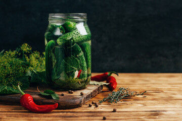 close-up on a jar of pickled cucumbers with greens seasonings with pepper on a wooden table on a dark background
