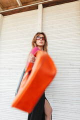 Stylish beautiful vogue model woman in fashion summer clothes with cool sunglasses and trendy orange bag posing in motion near a white vintage wooden building on the beach
