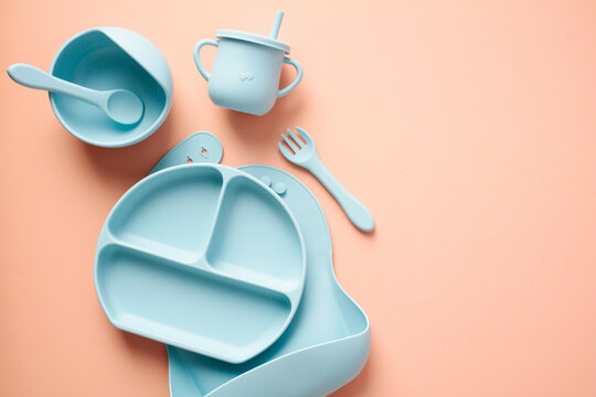 Baby feeding equipment. Flat lay silicone bib, plate, cup, cutlery on pastel beige background. Toddler self feeding training, baby weaning concept.