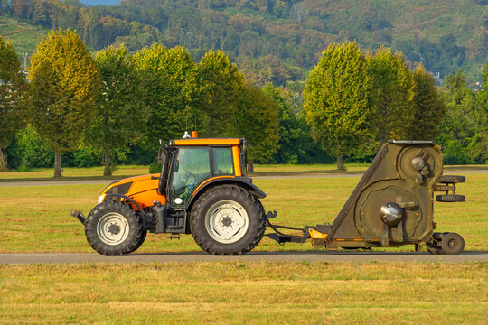 Yellow tractor with an industrial lawn mower trailer drives along the airfield at the airport, against the background of trees.
