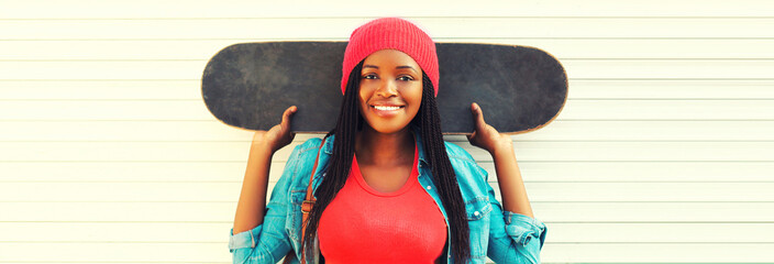 Portrait of happy smiling young african woman with skateboard wearing pink hat in the city