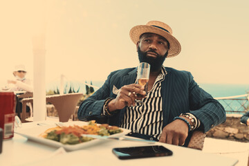 A portrait of an elegant mature bearded African man in a straw hat and a summer suit, having a break in a coastal restaurant after walking on a warm sunny day, sitting with a glass of lemonade