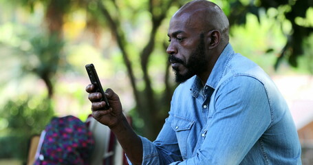 African man reading message on cellphone device outside at park, serious expression