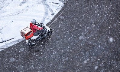 food delivery courier rides a black motorcycle under the snowfall with a yellow backpack. View from above