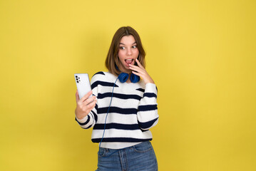 Funny girl holding mobile phone waiting for messages and congratulations from friends. Young smiling teenage  wearing earphones holding smartphone over yellow background. Copy space