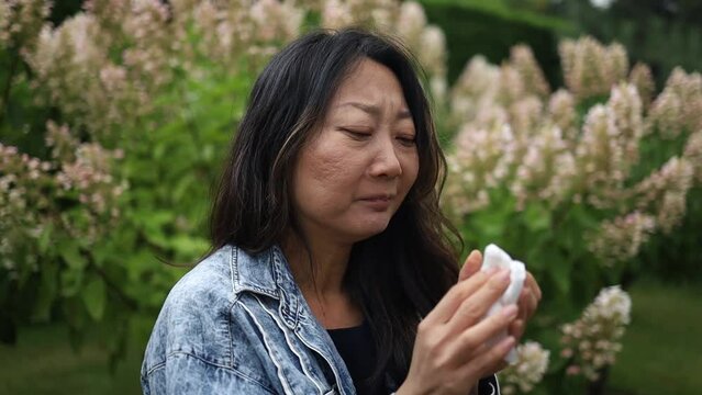 Sneezing young asian girl with nose wiper among flowering trees in the park
