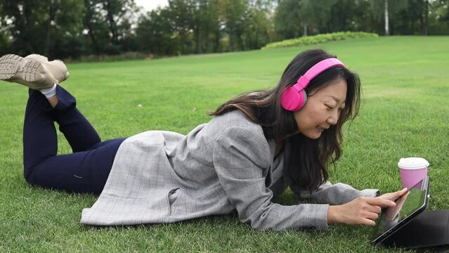 Young asian woman with digital tablet and headphone sits on the grass having conversation online in the park.