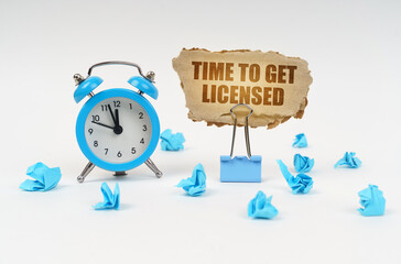 On a white surface there is an alarm clock, crumpled paper and a cardboard sign with the text - Time to Get Licensed