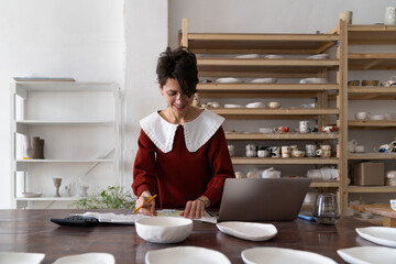 Smiling ceramic shop owner calculating business costs while working in pottery shop, female artisan making analysis of pottery studio expenses, doing bookeeping, working with invoices and receipts