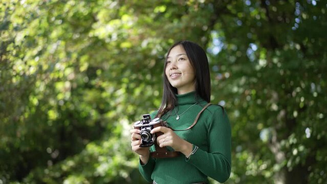 Portrait of confident talented Asian female photographer standing with camera in summer park smiling looking away. Medium shot of young slim woman enjoying hobby outdoors on sunny day. Slow motion