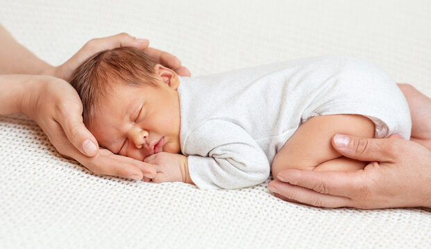 Newborn In Family Hands. Baby Sleeping In Mother And Father Palms Over White Blanket. Parents Holding Dreaming Infant. Cute Little Child Face Portrait. Childbirth And Parenting