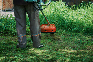 A worker mows grass with an electric or petrol trimmer on a lawn near a residential building. View...