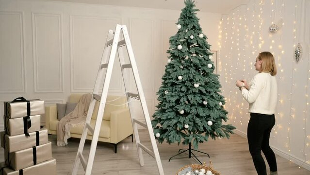 Woman in light clothes decorates a big Christmas tree standing on a ladder in the living room time lapse Christmas garlands and gifts.
