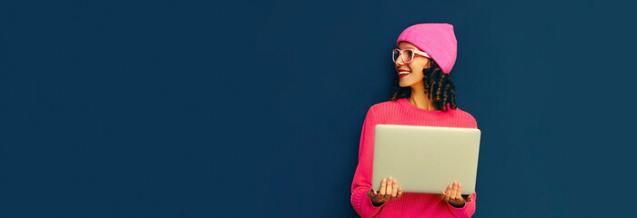 Portrait of stylish modern young woman working with laptop wearing pink colorful clothes on dark blue background, banner blank copy space for advertising text