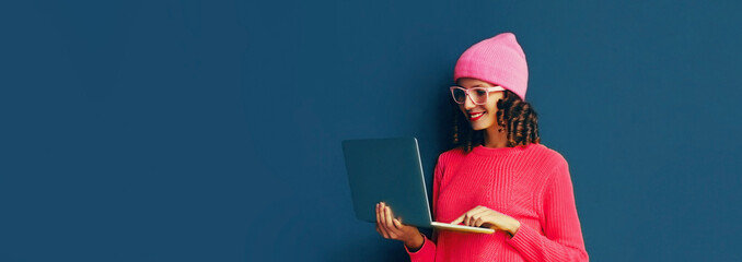 Portrait of stylish modern young woman working with laptop wearing pink colorful clothes on dark...