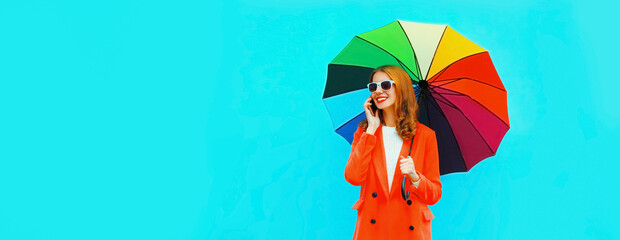 Portrait of happy smiling young woman calling on smartphone with colorful umbrella on blue...
