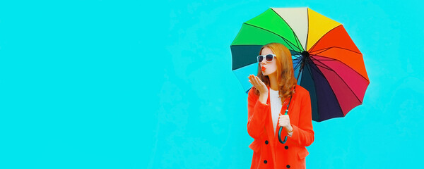 Autumn portrait of young woman with colorful umbrella blowing her lips sending air kiss wearing red jacket on blue background