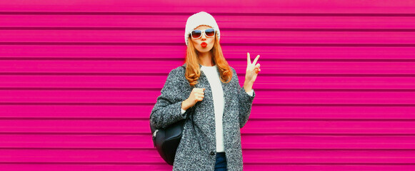 Portrait of stylish young woman blowing her lips sending air kiss wearing gray coat on colorful pink background