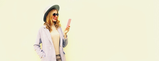 Portrait of stylish smiling woman taking selfie with smartphone wearing round hat, coat jacket on white background