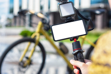 A male blogger holds a tripod in his hands with a smartphone mockup with a white screen and a light with a microphone on the background of a bicycle.