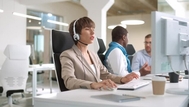Pretty professional Caucasian female worker talking in headset furniture while sitting in cabinet at desk and browsing on computer. Corporate worker. IT support job. Business concept. Close up