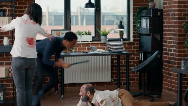 Afraid businessman hiding under table from hungry brain-eating monsters. Spooky evil monsters with deep and bloody wounds chasing apocalypse survivor in office while acting bizarre.
