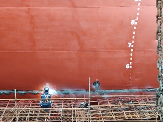 A red ship hull with draft mark and a man or worker who's sitting on the staging and welding on...