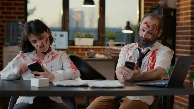 Brainless stupid office zombies sitting at desk while using smartphones. Spooky evil brain-eating monsters with deep and bloody wounds sitting in company workspace while using touchscreen devices.