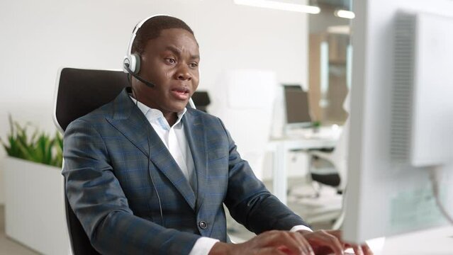 Handsome happy businessman sitting in modern cabinet and looking at monitor typing on computer keyboard and speaking in headset at workplace. Close up of African American man working on computer