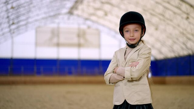 Young confident horse rider in black jockey helmet stands holding hands folded on her chest. Girl touchй her hat looking at camera. Close up. Blurred backdrop.