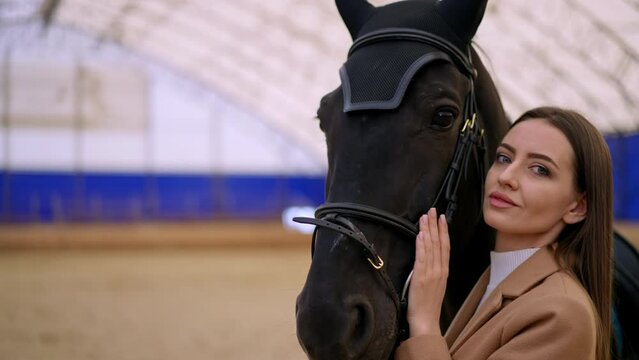 Mid-aged young woman with long dark hair holding a horse by the bridle. Close up. Woman caressing the animal before riding it.