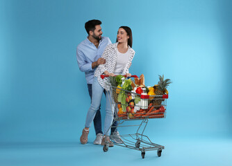 Young couple with shopping cart full of groceries on light blue background © New Africa