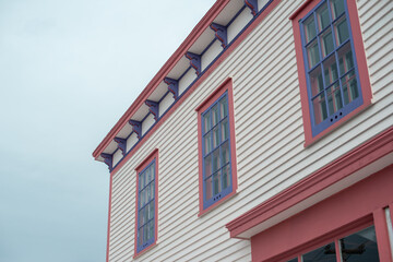 The roof section of a large white vintage wooden building with decorative pink and purple wood trim. There are three multi-pane windows on the top floor. The background is a dramatic blue cloudy sky.