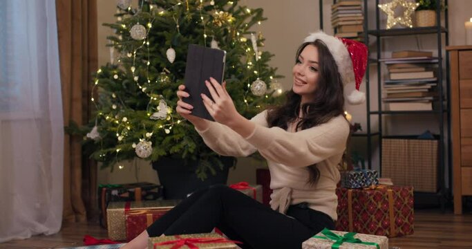 A young attractive girl is sitting on floor near Christmas tree and has video chat on tablet with friend. The girl communicates, smiles and shows the gift she received. There are presents on floor.