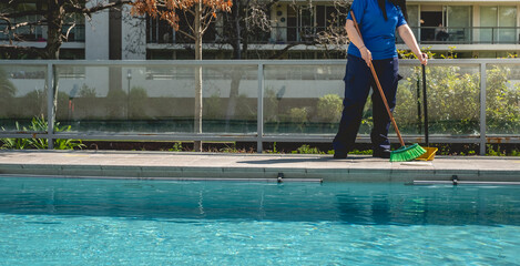 Young latino janitorial worker lady in blue uniform sweeping and cleaning at the side of big swimming pool