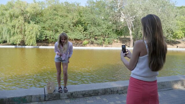 a girl takes pictures of her friend on the phone near the water in the park, who dances for tiktok