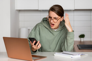 A young woman is sitting in the kitchen in front of a laptop and and reads the message on the phone. On the face of a woman, the emotion of bewilderment of surprise or disappointment