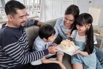 Happy Asian Thai family, young son is surprised with birthday cake, blows out candle, and celebrates joy party with parents and sister in living room together, wellbeing domestic home special event.