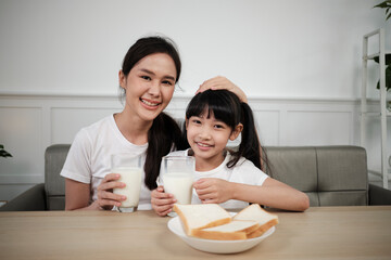 Obraz na płótnie Canvas Portrait of healthy Asian Thai family, happy daughter, and young mother looking at camera, drink fresh milk and bread together at dining table in morning, wellness nutrition breakfast meal lifestyle.