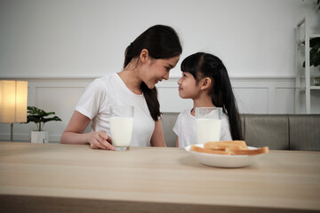 Obraz na płótnie Canvas Healthy Asian Thai family, happy little daughter, and young mother face to face at dining table with fresh milk in glass and bread care together, wellness nutrition breakfast meal, morning lifestyle.