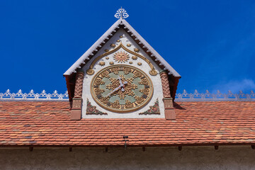 Beautiful patterned clock on a tower with a red tiled roof against a blue cloudless sky. The dial with Roman numerals is intricately decorated.