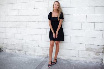 Beautiful natural blond woman with long hairs  walking in the street . Girl dressed black  dress and posing against street wall